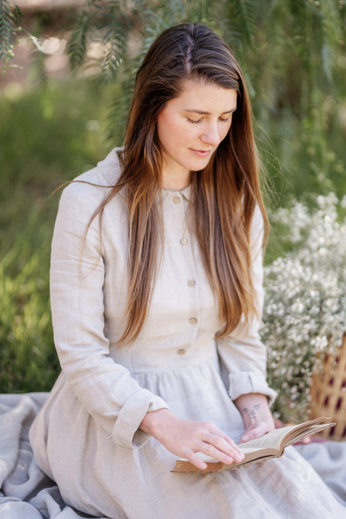 Woman with longer brown hair sitting in nature and reading a book. She is wearing a long-sleeved natural linen dress that has a Peter Pan collar and wooden buttons that go from the neckline to the waist