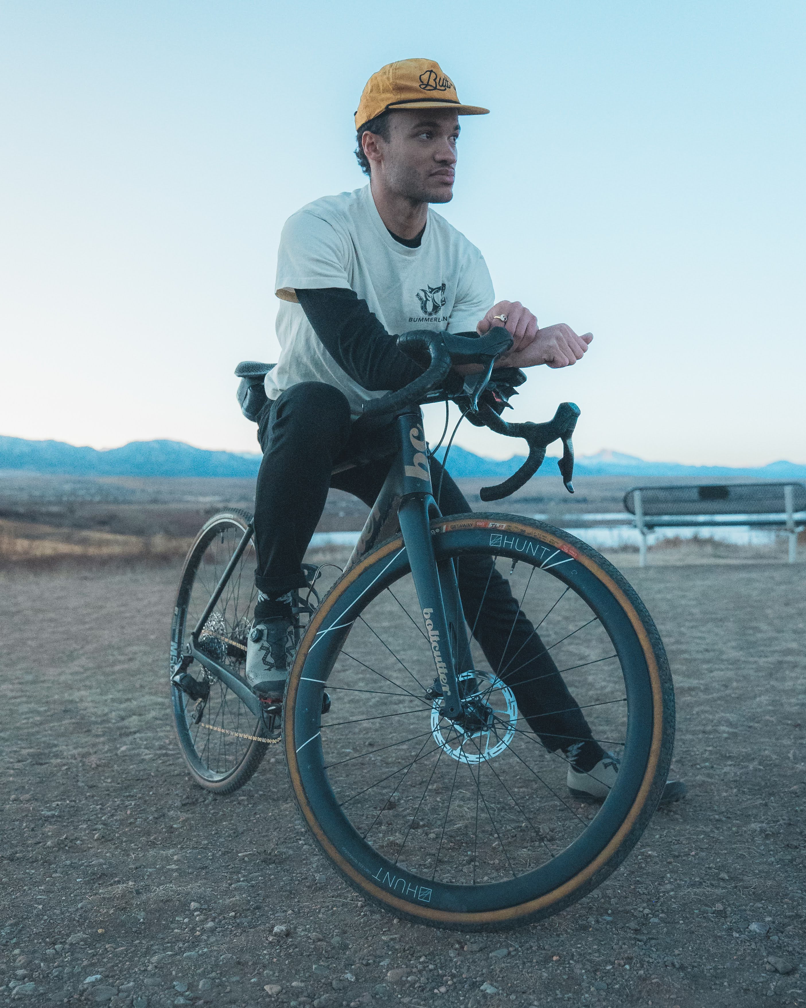 Person sitting on a bike outdoors, wearing a yellow cap and casual attire.