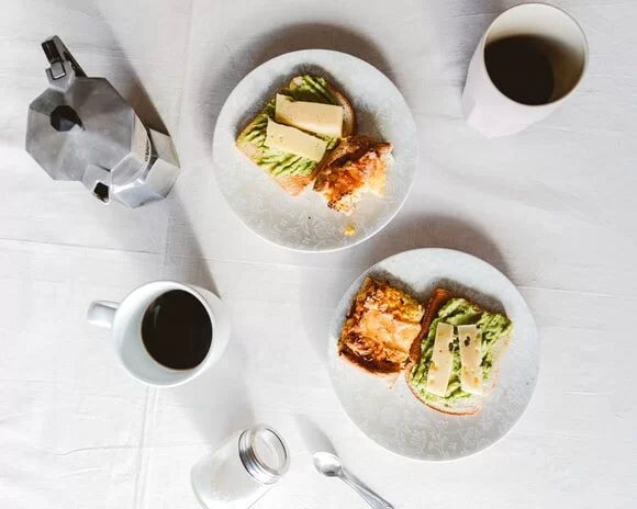 Two plates with avocado toast and cheese beside mugs of black coffee—an intermittent fasting-friendly breakfast that supports energy, satiety, and longevity when paired with healthy fats and antioxidant-rich coffee.