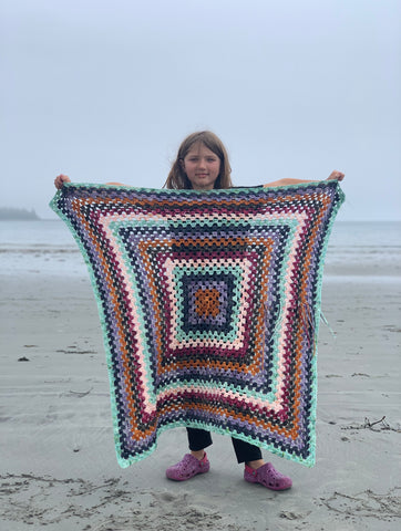 a cute kiddo at the beach in Maine on a foggy day,  holding up a large granny square blanket made from leftover yarn