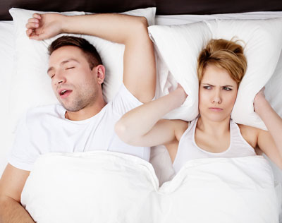 photo of a man and woman lying in bed. a man is sleeping, and the woman next to him is covering her ears with a pillow, looking disgruntled