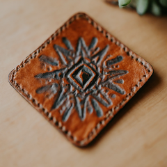 Four square leather coasters in different colors and textures, displayed on a wooden table with water droplets on them.