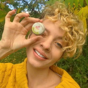 Beautiful smiling woman holding a lip balm tin in front of her right eye.