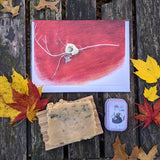Bird nest on twig cards, botanical soap, and soothing salve lay flat on a weathered table with red and yellow fall leaves around them.