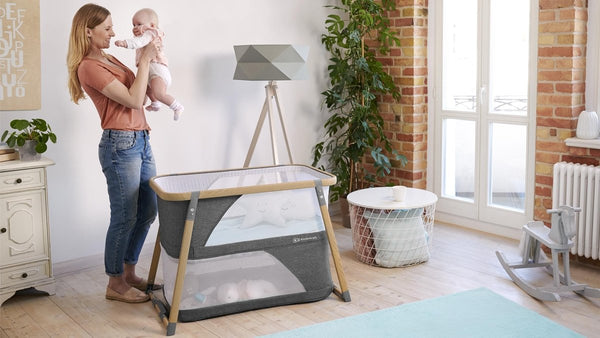 Mother holding baby next to Kinderkraft Baby Travel Cot SOFI PLUS