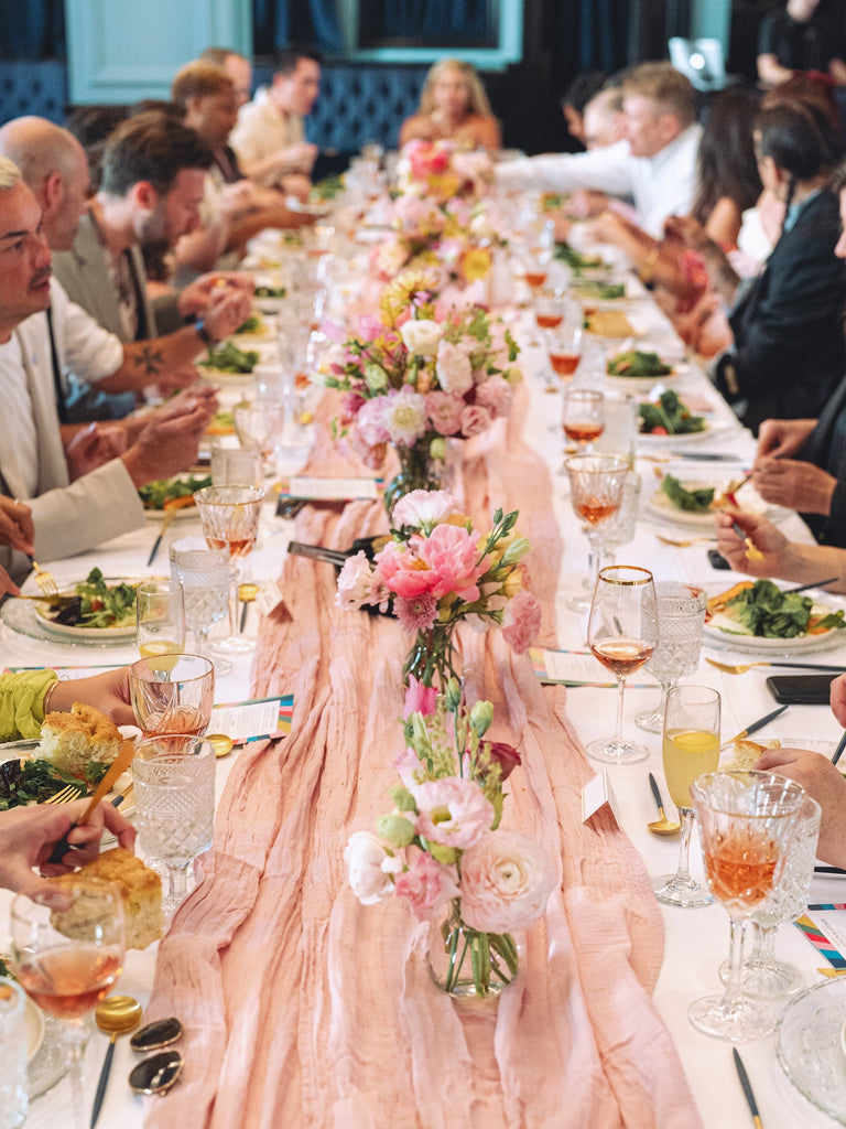 A full bright dining table with flower arrangements, plates of food, wine glasses, and guests eating