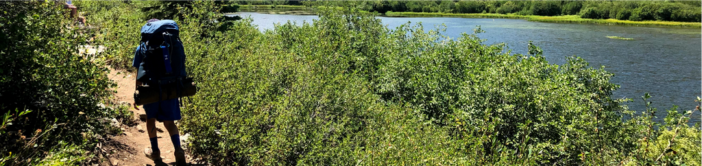 One of Susan's sons hiking with his pack along the trail of a lake.