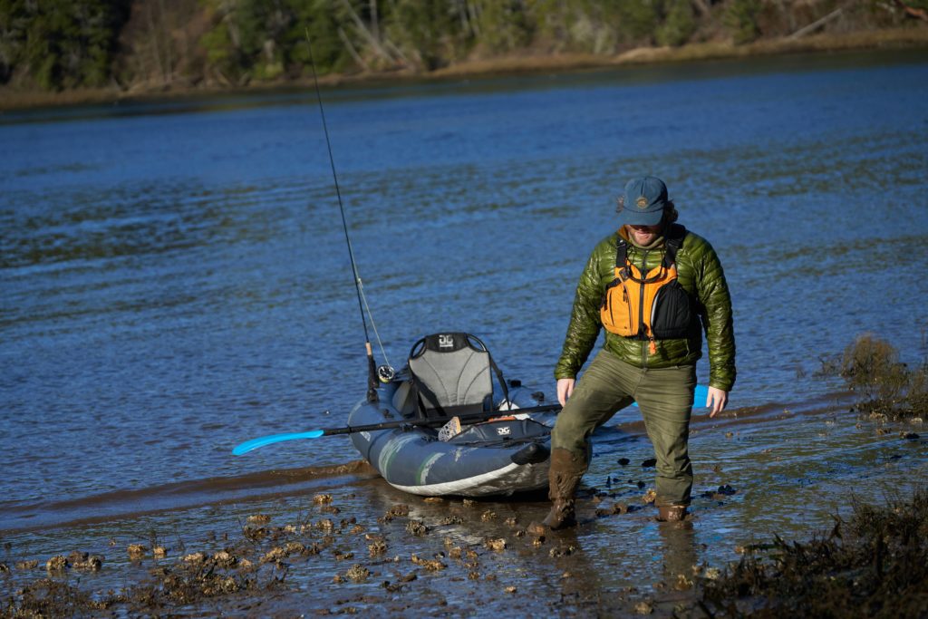 A paddler standing in mud up to his knees while pulling in inflatable fishing kayak up a beach