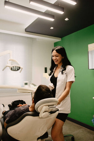 Female dental hygienist in lab attire standing over male patient in chair in front of a green wall