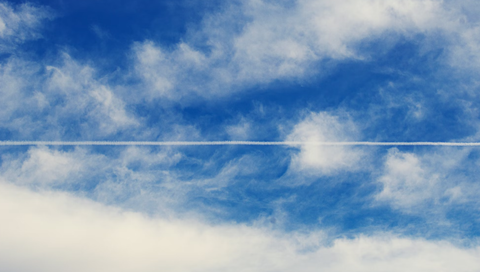 A white contrail streaks across a vibrant blue sky, leaving a trail through fluffy white clouds.