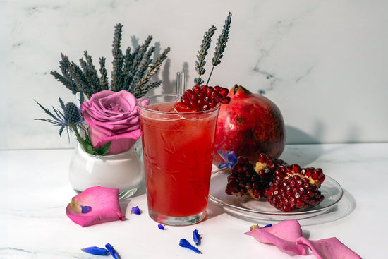 French Kiss lavender pomegranate gin cocktail in a  white marble scene with dried lavender spikes, fresh pomegranate fruits, a purple rose, and scattered petals.