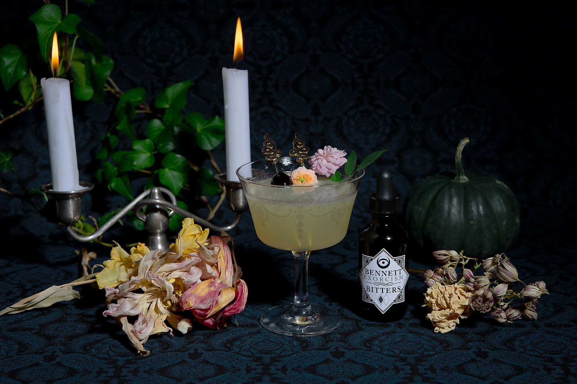 Bottle of Exorcism Bitters next to Corpse Reviver #2  cocktail in a moody setting with candles, a dark green pumpkin, and botanicals.