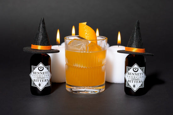 Orange colored Witch Hat cocktail in a rocks glass next to bottles of Exorcism Bitters that have little witch hats on them, with candles in the background.