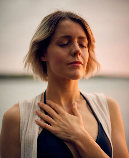 A blond woman in a black bikini top and white vest closes her eyes and places her hand on her chest. Image by Darius Bashar