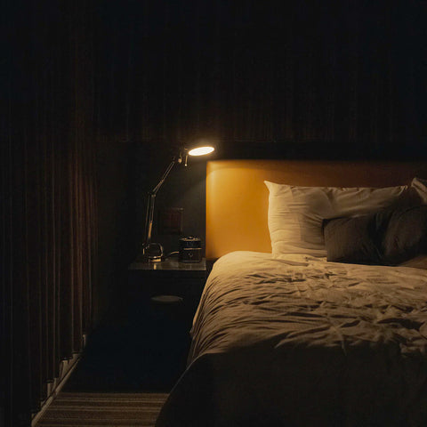 A nightstand reading lamp illuminates a tan headboard and bed with white sheets in a darkened room. Image by JP Valery, Unsplash.