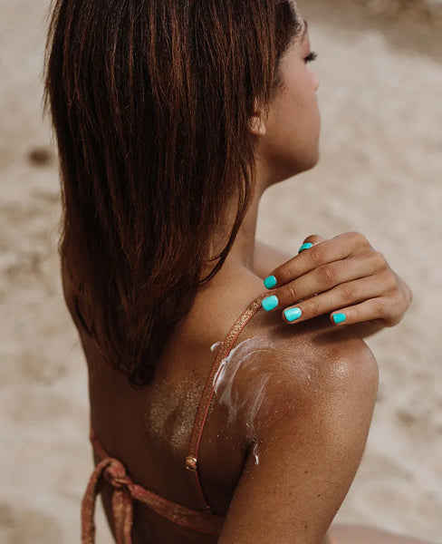 A woman puts sunblock on her back while wearing a bikini and sitting on the beach. Image by Karolina Grabowska