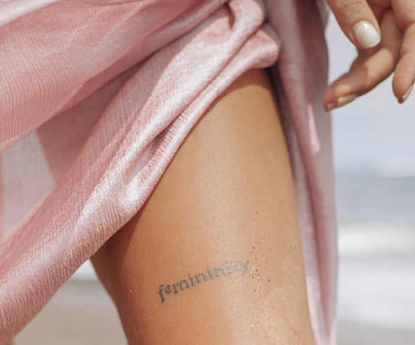 A woman with the word femininity tattooed on her thigh walks on the beach in a pink coverup. Image by Ksu&Eli.