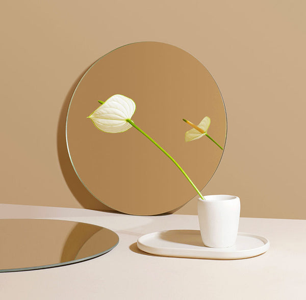 A white Anthurium flower sits in a white cup on a white tray and is reflected in a round mirror in front of a tan wall. Image by Marina Podrez.