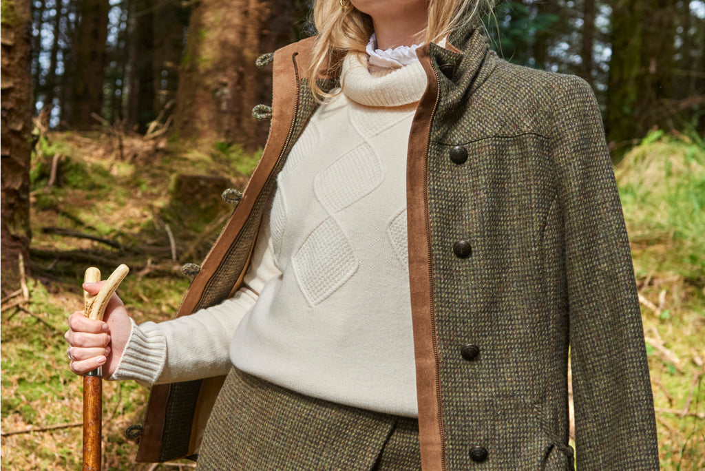 Woman in a forest wearing a white roll-neck sweater or jumper and a green tweed jacket.