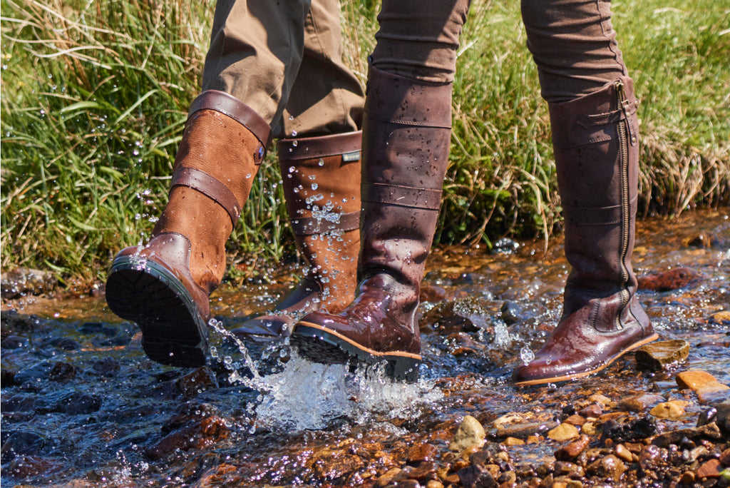 The brown Roundstone leather waterproof and breathable boot is walking through a stream with rocks at the bottom and some vegetation in the background.