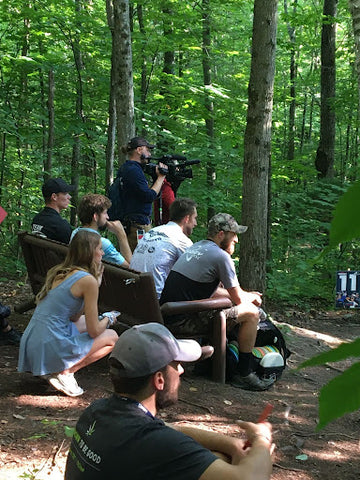 Spectators sitting lined up on a bench behind a tee watching a disc golf tournament.