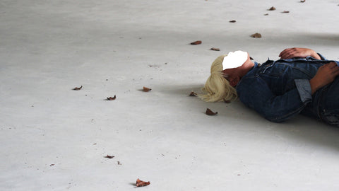 A person in a jean jacket and short blonde wig lying on the ground with fallen leaves. Their face is cut out in white.