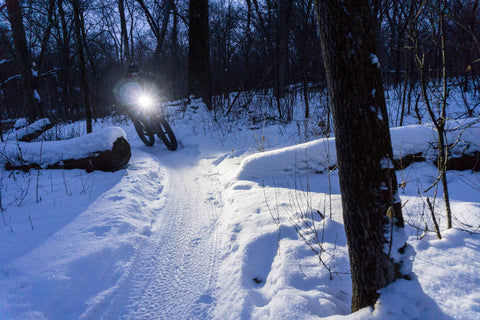 A cyclist rides around a curve on a snowy trail with the headlight shining.