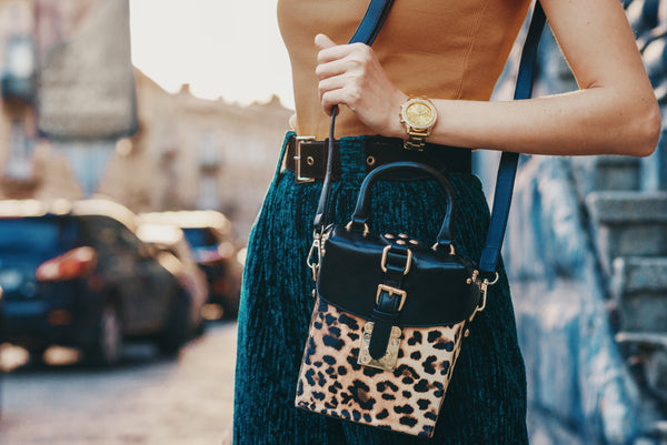 A cropped image of a woman wearing an animal print cross body bag. She also has highwaisted dark teal trousers and a cinnamon colored top.