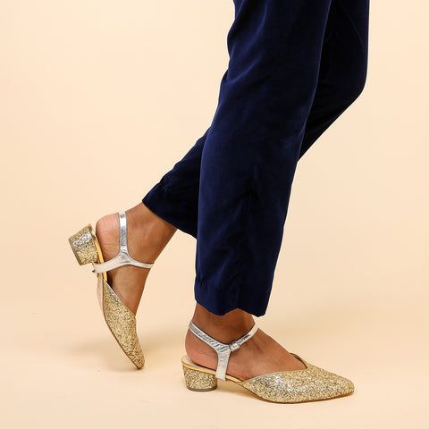 Alterre's new holiday shoes, the Gatsby Glitter V Slide + Silver Jackie show cases layers of chunky glitter in muted golds.  A women is walking in them wearing blue velvet dress pants.