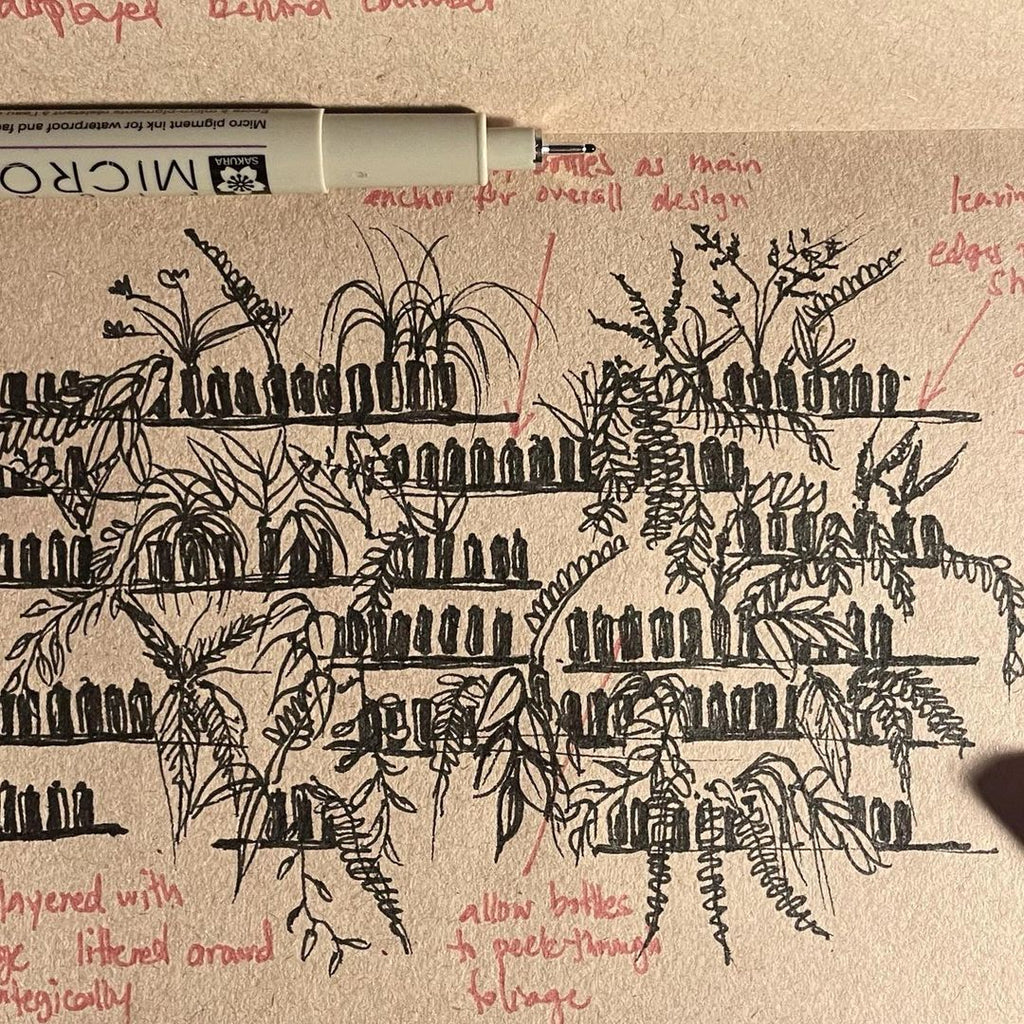 A hand-drawn sketch on brown paper, possibly a design concept, featuring multiple shelves stocked with bottles, flanked by plants with various leaf patterns. The sketch is annotated with red ink, highlighting design elements such as allowing bottles to peek through foliage and noting the plants as the main anchor of the overall design. A pen rests on the page, indicating an ongoing design process.