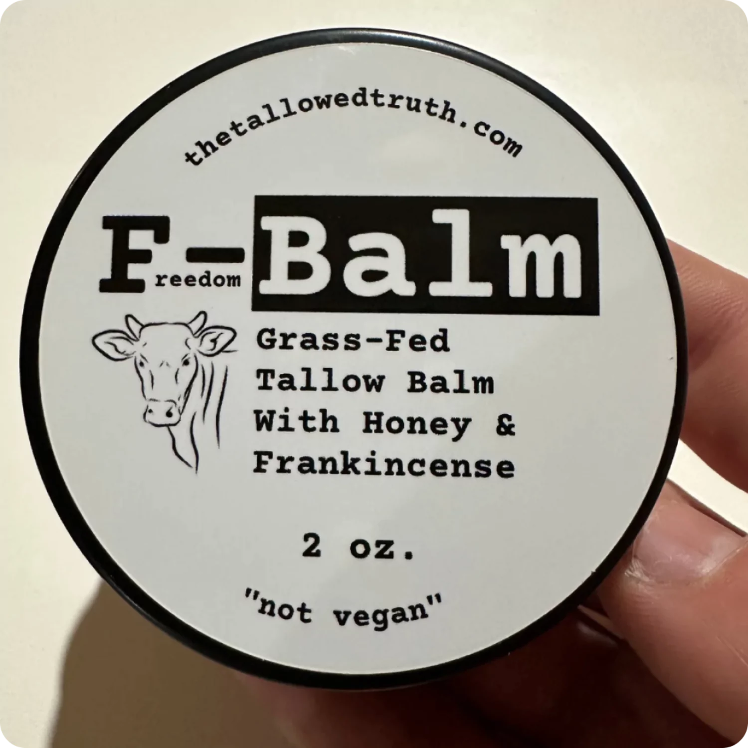 'Hand holding a 2 oz. tin of grass-fed tallow balm with honey and frankincense.'