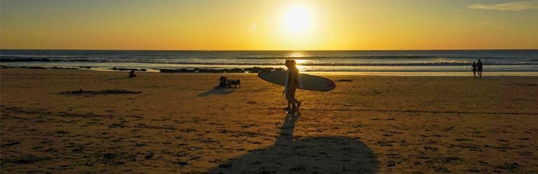 A person walks across a beach in Costa Rica holding a surfboard. The Sun is setting behind them and the whole sky is gold.