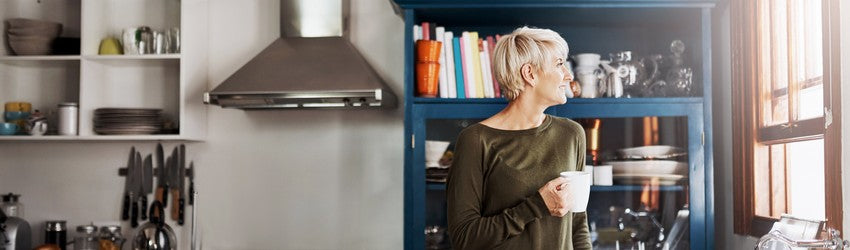 Blonde woman standing and drinking coffee in a modern blue kitchen while looking out the window which is to the left of the image.