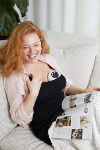 Mum laughing relaxed on couch pumping with hands free wearable breast pump melony whilst reading a magazine