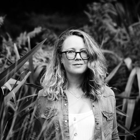 Black and white image of ALP Ceramics founder Adrienne Pitts. She is a pakeha woman wearing glasses standing amongst flax plants.