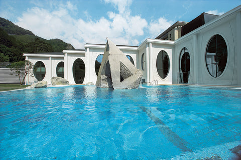 External view of a swimming pool with a big installation made by rock inside and a white building in the back
