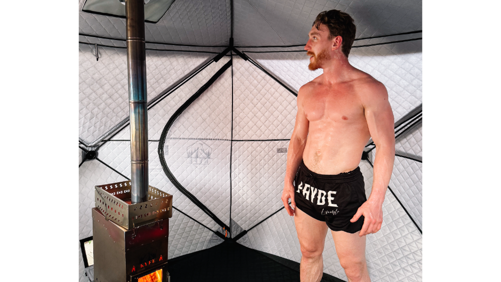 Man standing shirtless inside an insulated portable sauna tent, sweating beside a burning stove with a metal chimney.