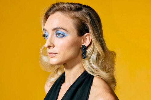 A young woman with blonde hair and blue eyeshadow wears zig zag shaped leather earrings.