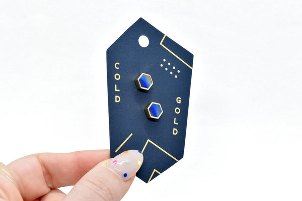hand holding a dark teal card with gold marks and cold gold logo, cards includes sapphire earrings with gold hexagon rim in 14k gold and sterling silver posts, 14k gold plated posts
