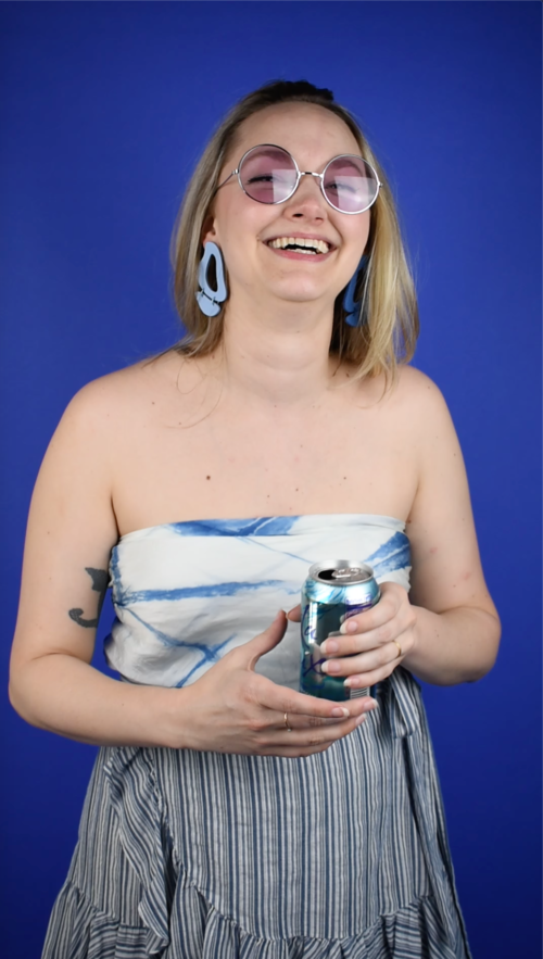Alaina holds a can of La Croix Pure while wearing cut out leather earrings in periwinkle with a scarf top and wrap skirt. 