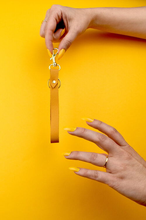 Una mano con uñas de color amarillo brillante muestra una pulsera de cuero auténtico de color amarillo caléndula con llaveros y cierres de oro.