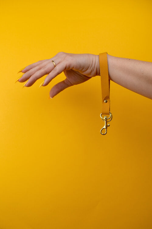 Una mano con uñas de color amarillo brillante muestra una pulsera de cuero auténtico de color amarillo caléndula con llaveros y cierres de oro.