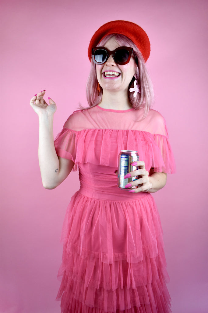 Shelby laughs over a can of Razz-Cranberry La Croix while wearing a pink party dress, a red beret, and a mismatched pair of Point Move earrings in red and metallic pink leather. 