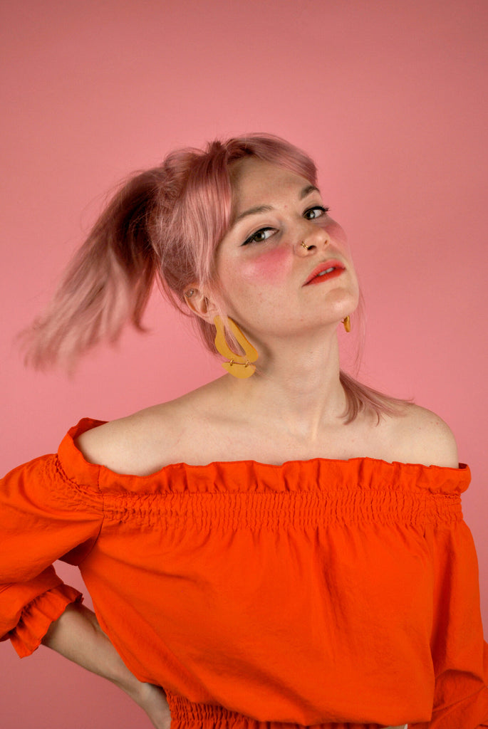 Shelby wears the Inferno earrings in Marigold yellow leather with an off-the-shoulder top in bright orange and finishes the look with bright pink makeup.