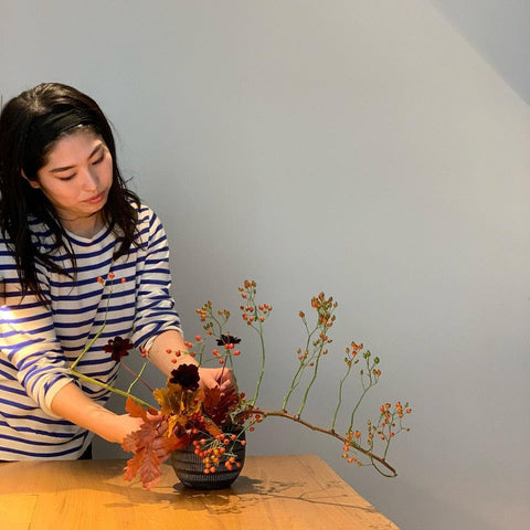 A woman in a striped top organises orange, gold and burgundy foliage into an Ikebana arrangement.