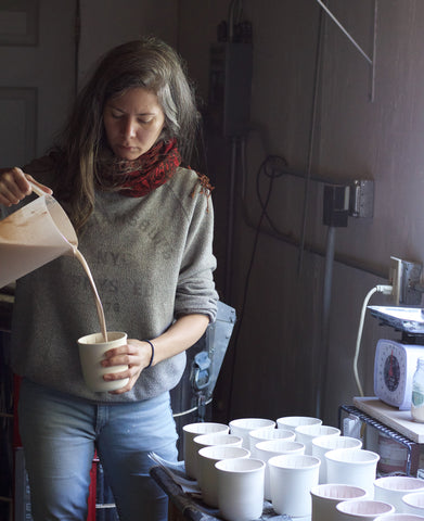 Artist pouring glaze into a piece from a pitcher with other pots waiting to be glazed on a side table