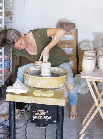 Woman poised throwing at a pottery wheel with freshly made cups on a side table