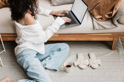 a woman with shoulder length black curly hair wearing a white shirt and light wash denim jeans. she's sitting on the floor on a laptop that's sitting on a coffee table