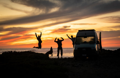 silhouette of three people and a dog jumping next to a van on the beach at sunset