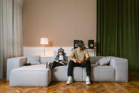 Two boys sit on a couch with VR headsets on while playing video games